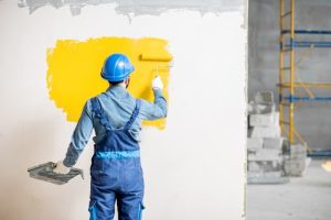 Workman in uniform painting wall with yellow paint at the construction site indoors